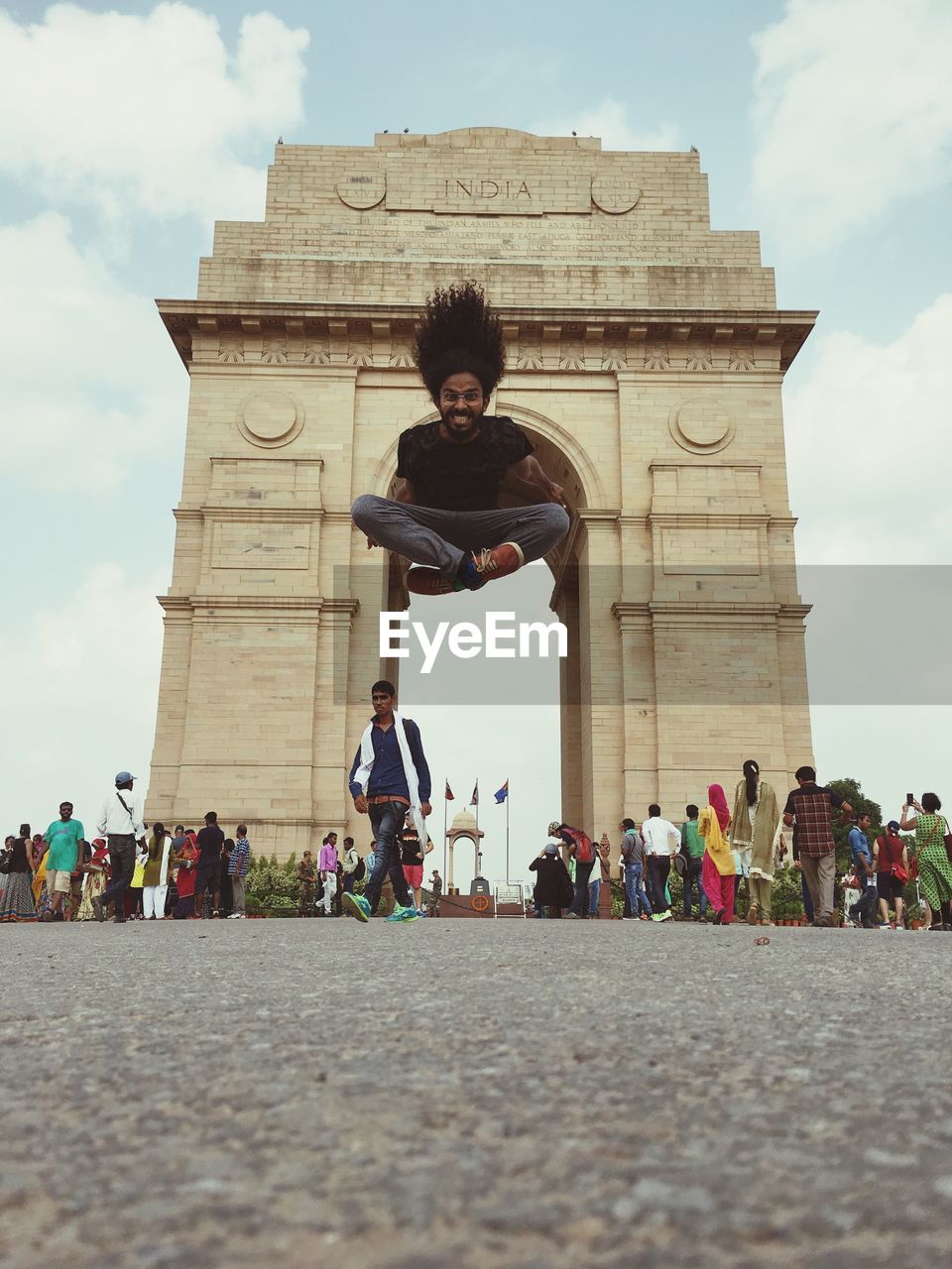 Young man jumping against india gate with people walking on street in city