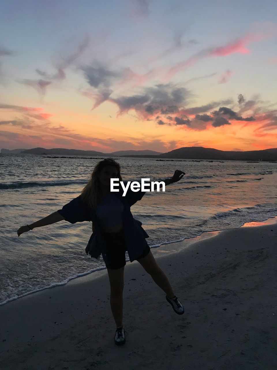 Portrait of young woman jumping at beach against sky during sunset