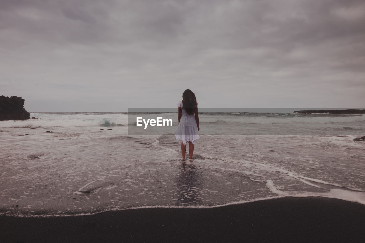 Rear view of woman standing at beach against cloudy sky