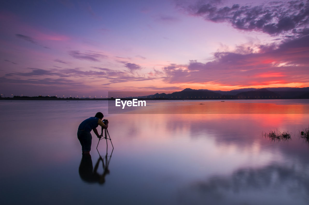Man photographing while standing in lake during sunset