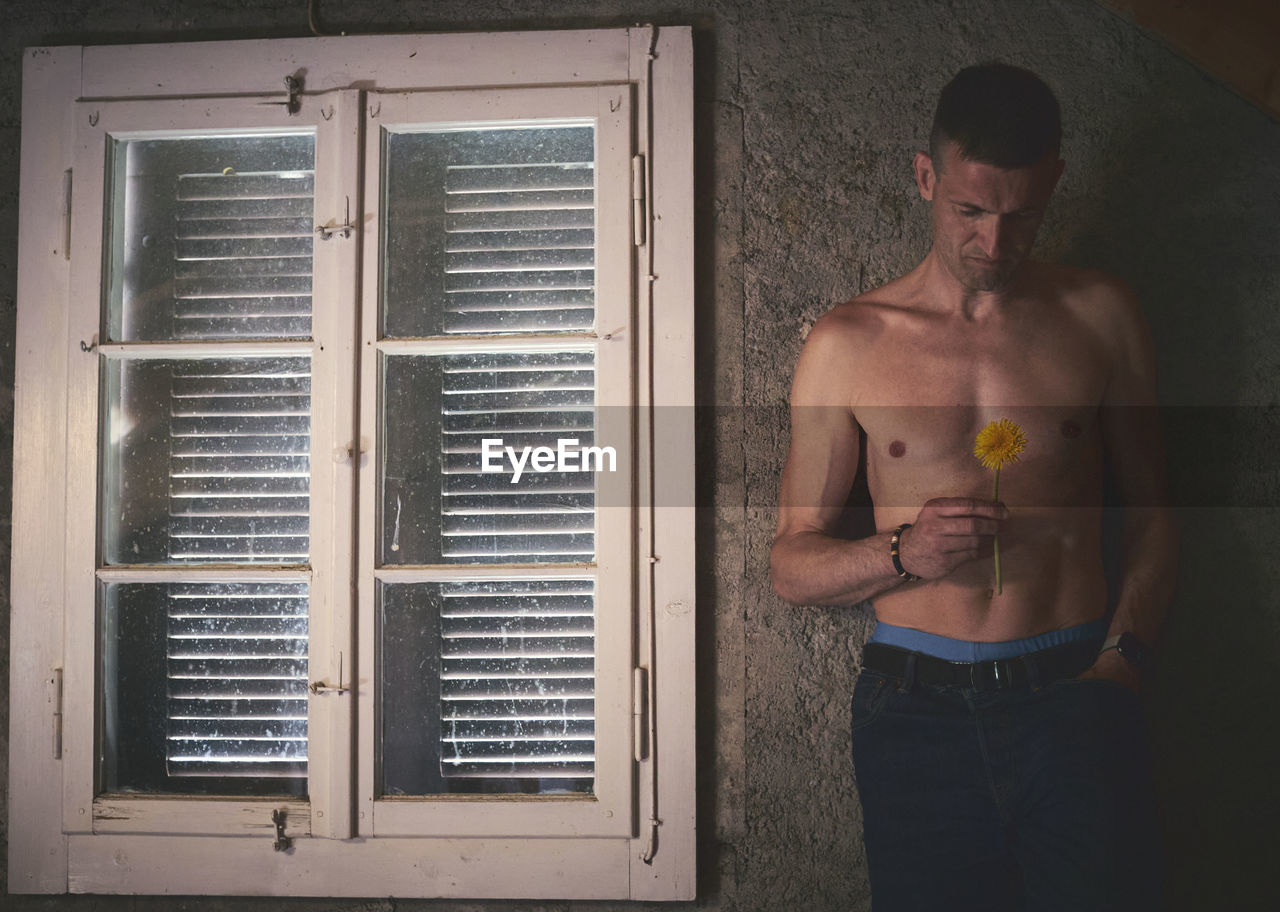 Full length of shirtless man standing by window and holding a flower