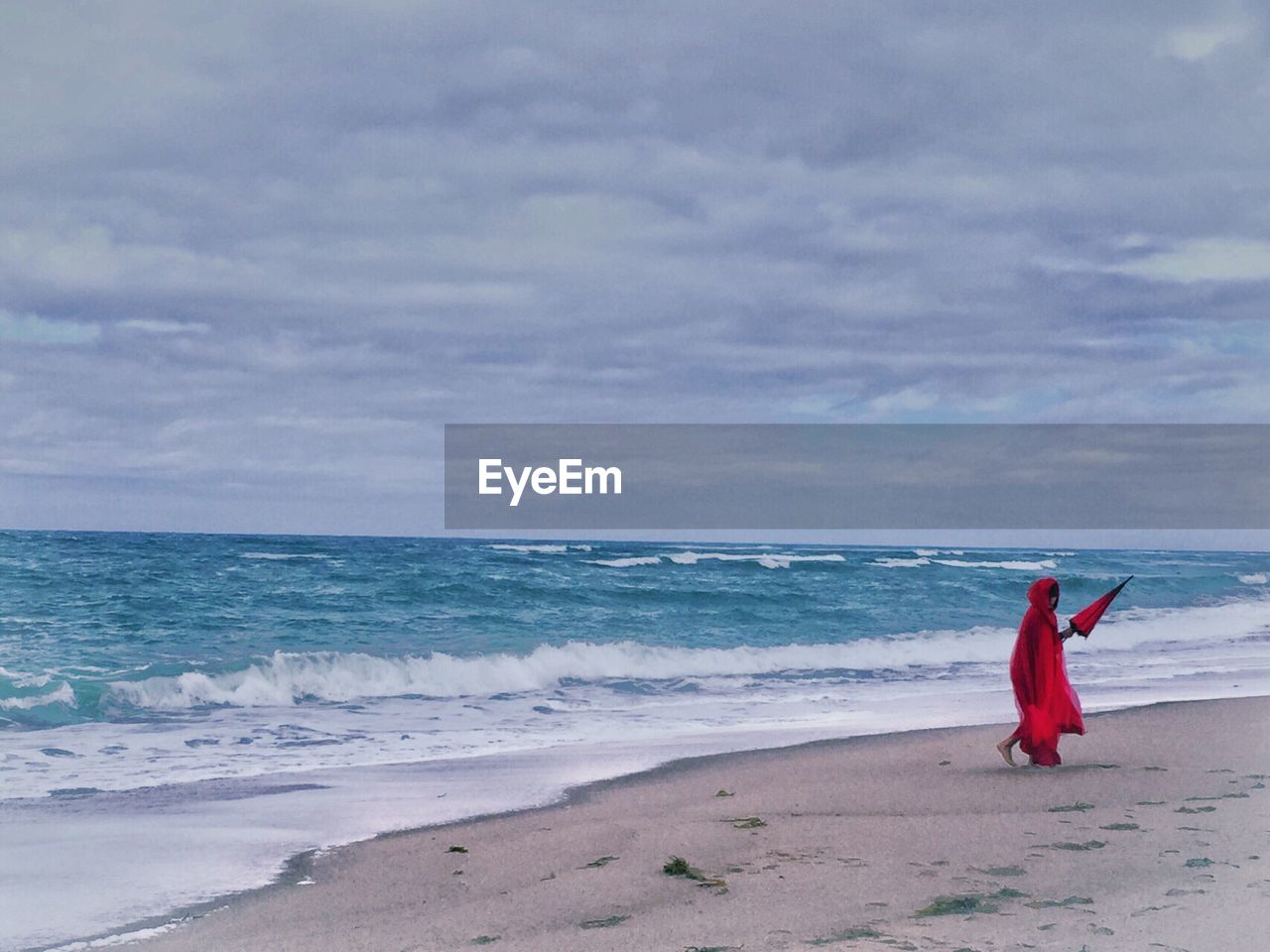 Person with red umbrella walking at beach against cloudy sky