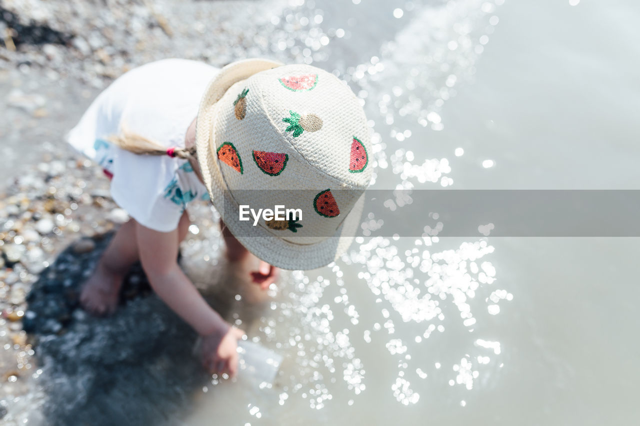 High angle view of girl wearing hat filling water in container on shore at beach during sunny day