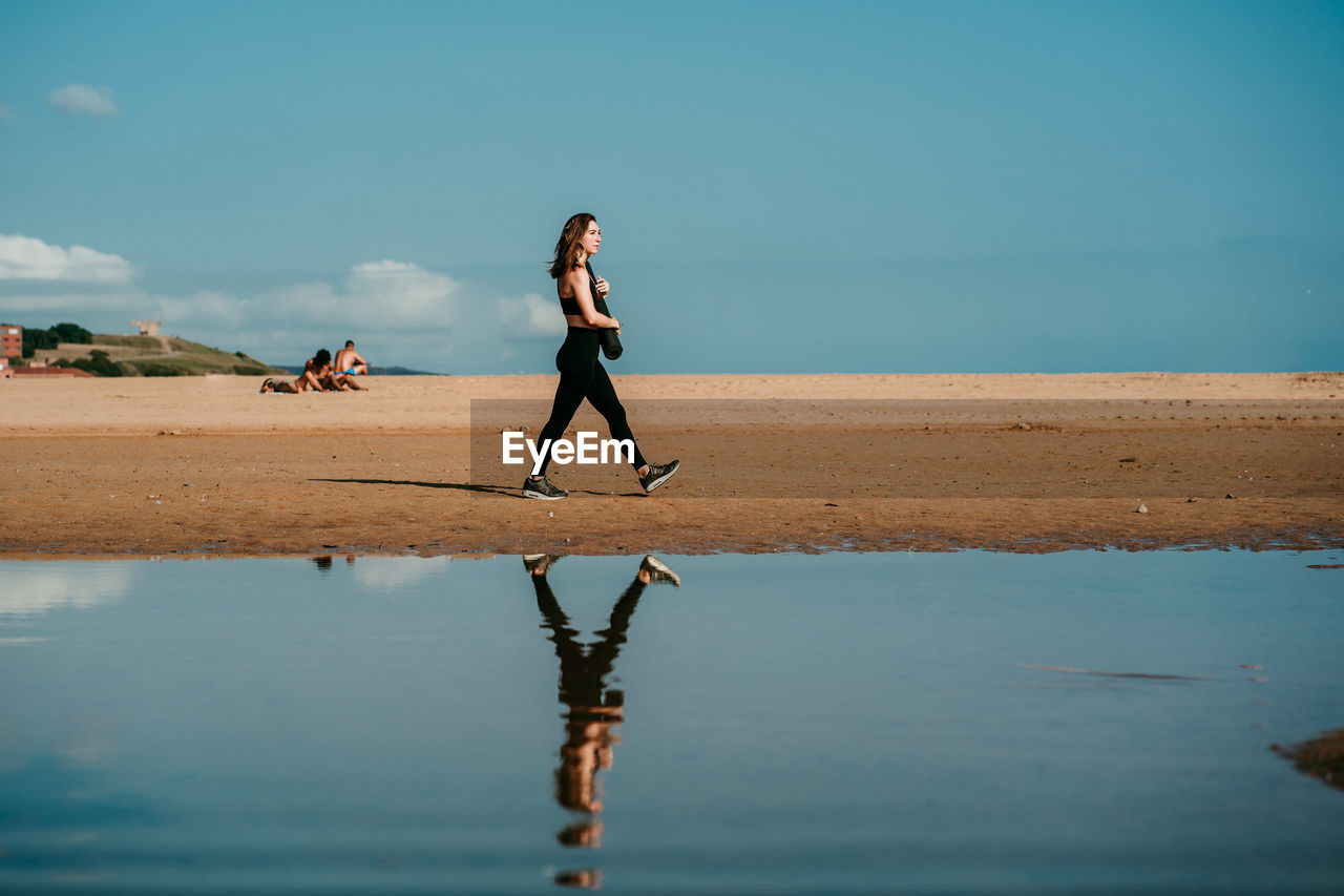 Side view of slim female in sportswear and with yoga mat walking along beach near river on sunny day