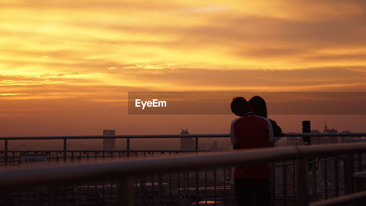 Rear view of silhouette couple standing by railing against orange sky during sunset