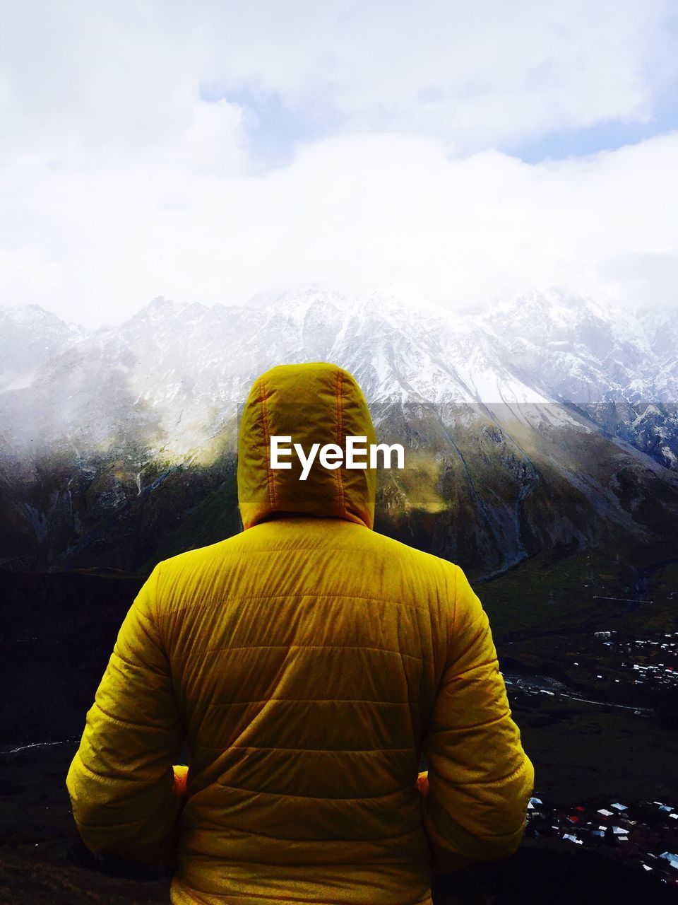 Rear view of person in yellow hooded jacket standing against snowcapped mountains