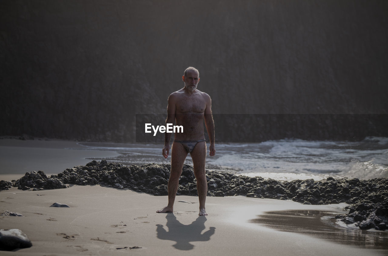 Full length of shirtless man  with swimwear standing on beach 