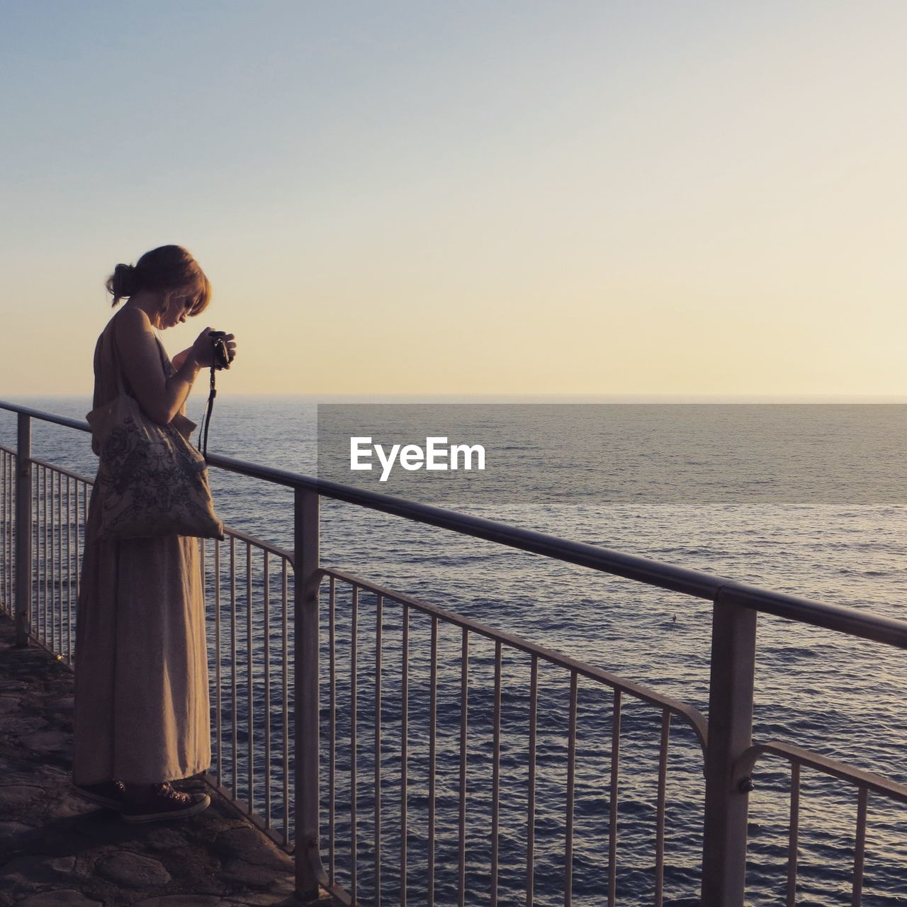 Full length of woman holding digital camera by sea against clear sky during sunset