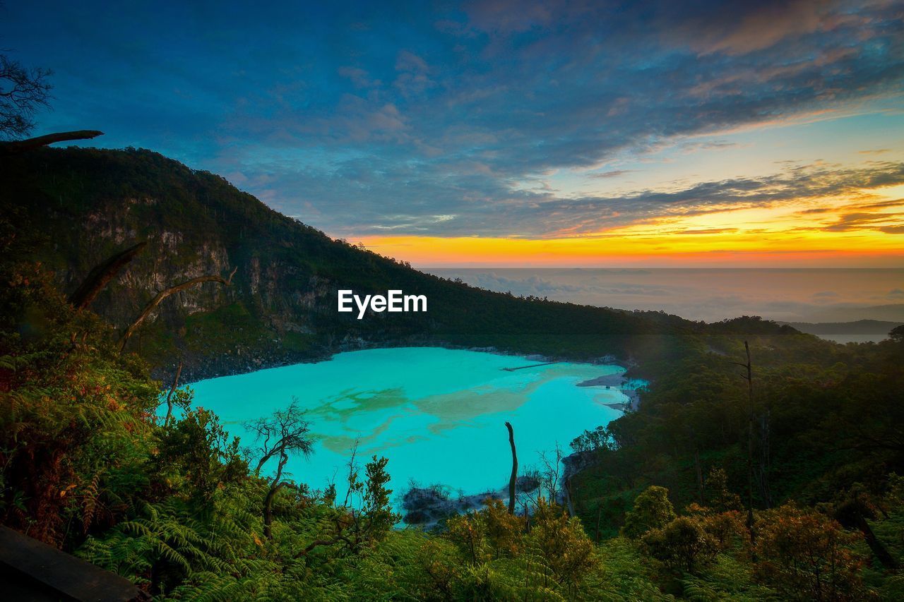 Scenic view of turquoise lake by mountains against dramatic sky during sunset