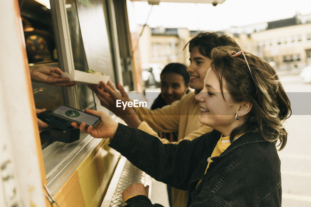 Smiling young woman doing online payment through smart phone while buying food from concession stand