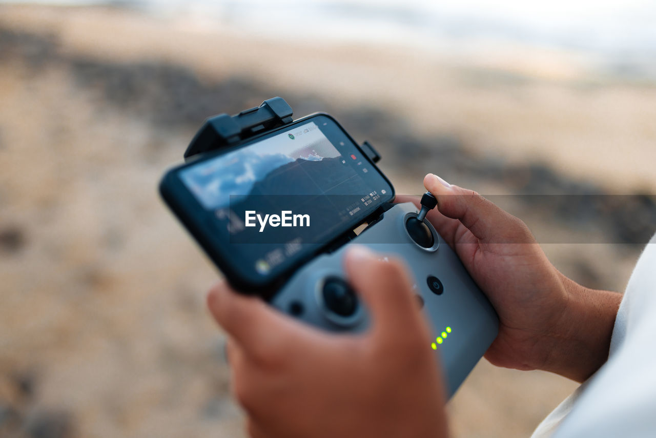 Soft focus of unrecognizable ethnic tourist using remote control and smartphone to fly drone over sea and shoot waves on weekend day on beach