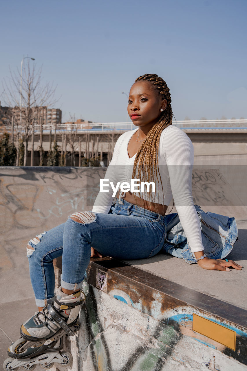 Cool black female with braided hairstyle and in rollerblades sitting on ramp in skate park and looking away