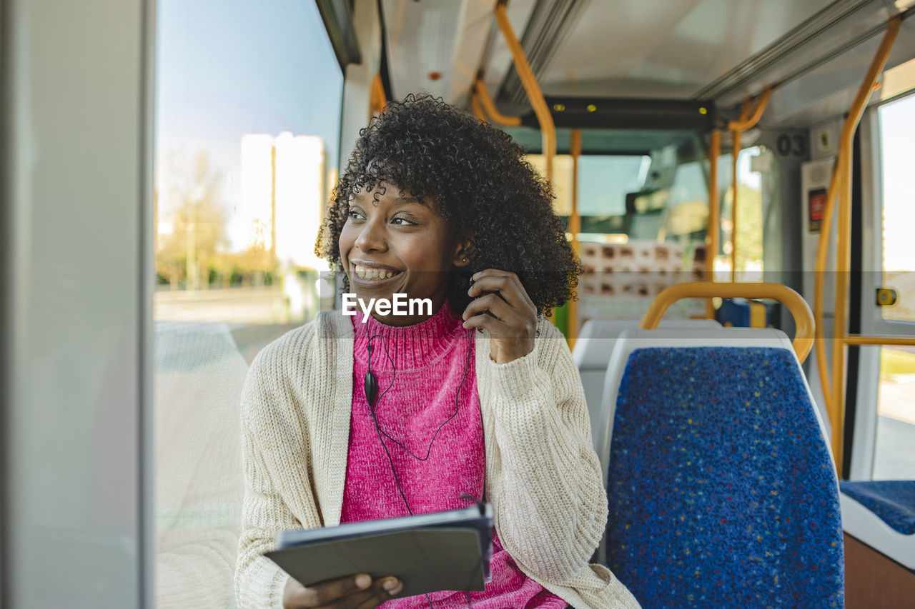 Happy young woman wearing in-ear headphones holding tablet pc in tram