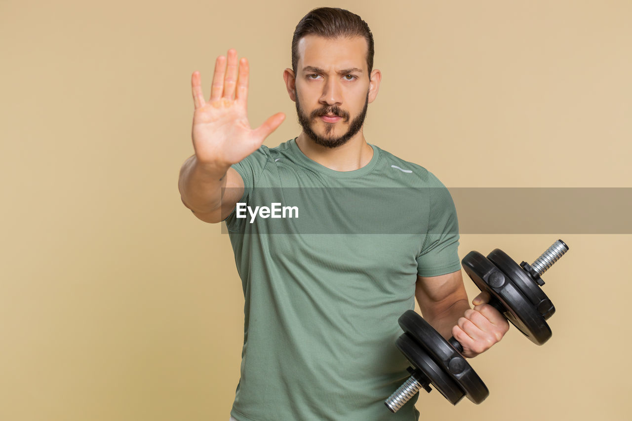 one person, adult, studio shot, men, indoors, portrait, colored background, hand, waist up, young adult, casual clothing, standing, holding, looking at camera, copy space, arm, front view, emotion, lifestyles, t-shirt, facial hair, finger, beard, cut out, communication, muscular build, strength, gesturing, sports
