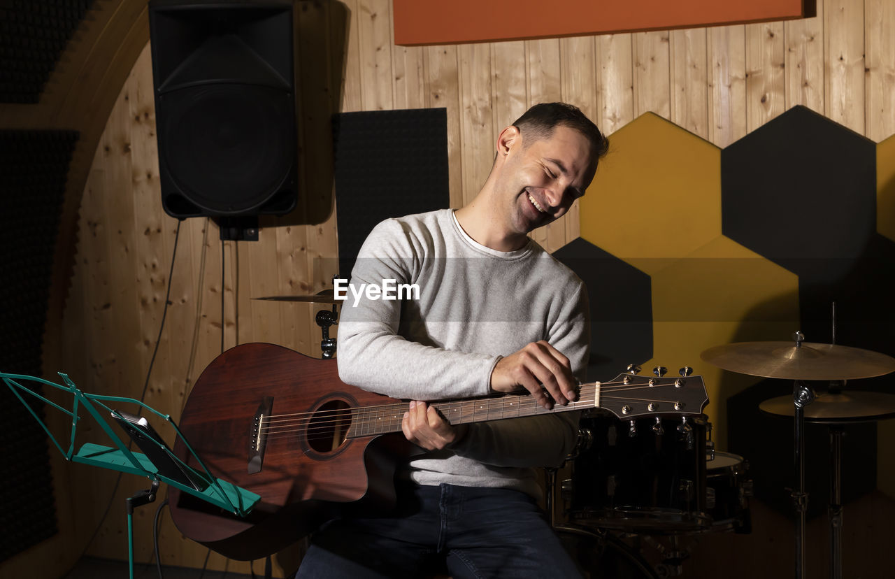 Smiling white man tuning guitar, playing, holding musical instrument in hands, sitting on chair in