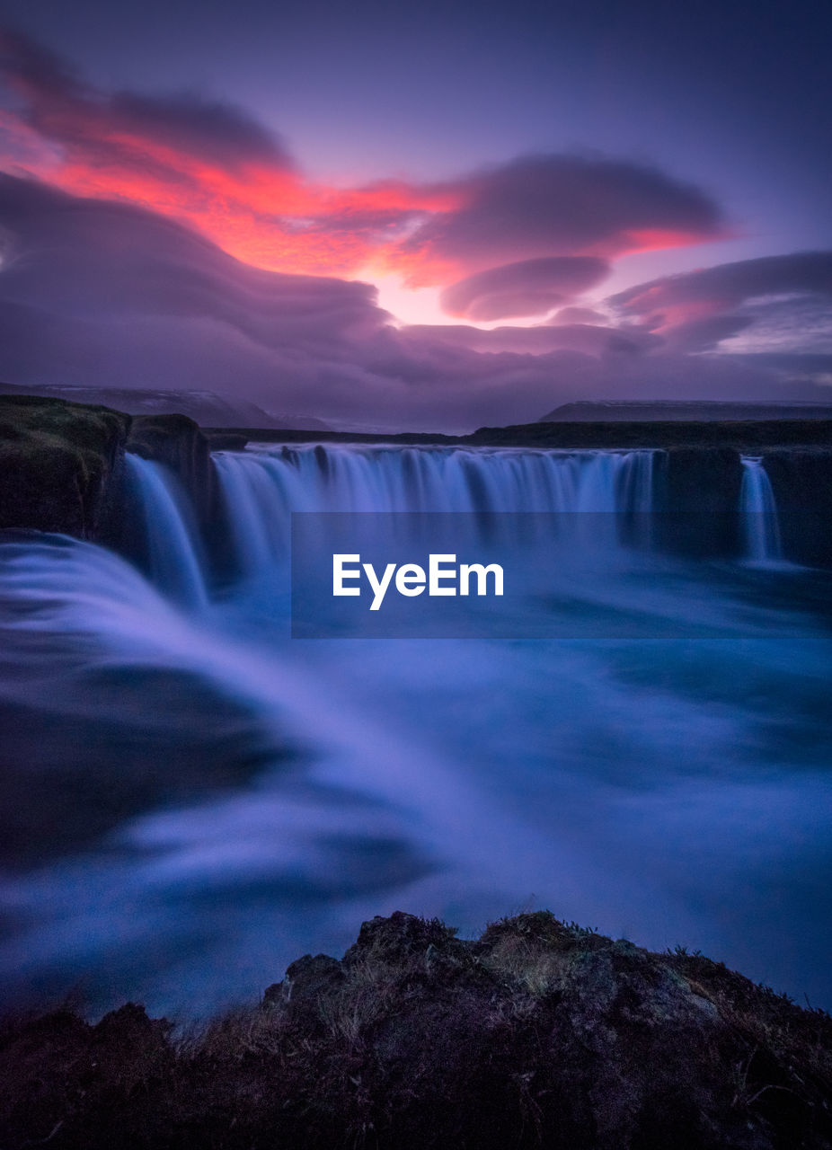 Scenic view of waterfall against dramatic sky during sunset