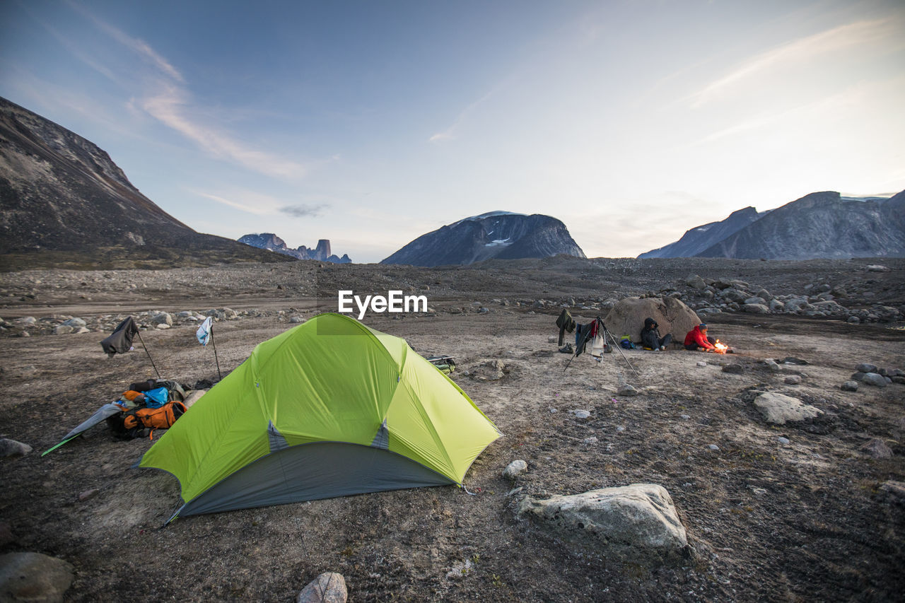 Two men prepare late dinner during dusk at arctic campsite.