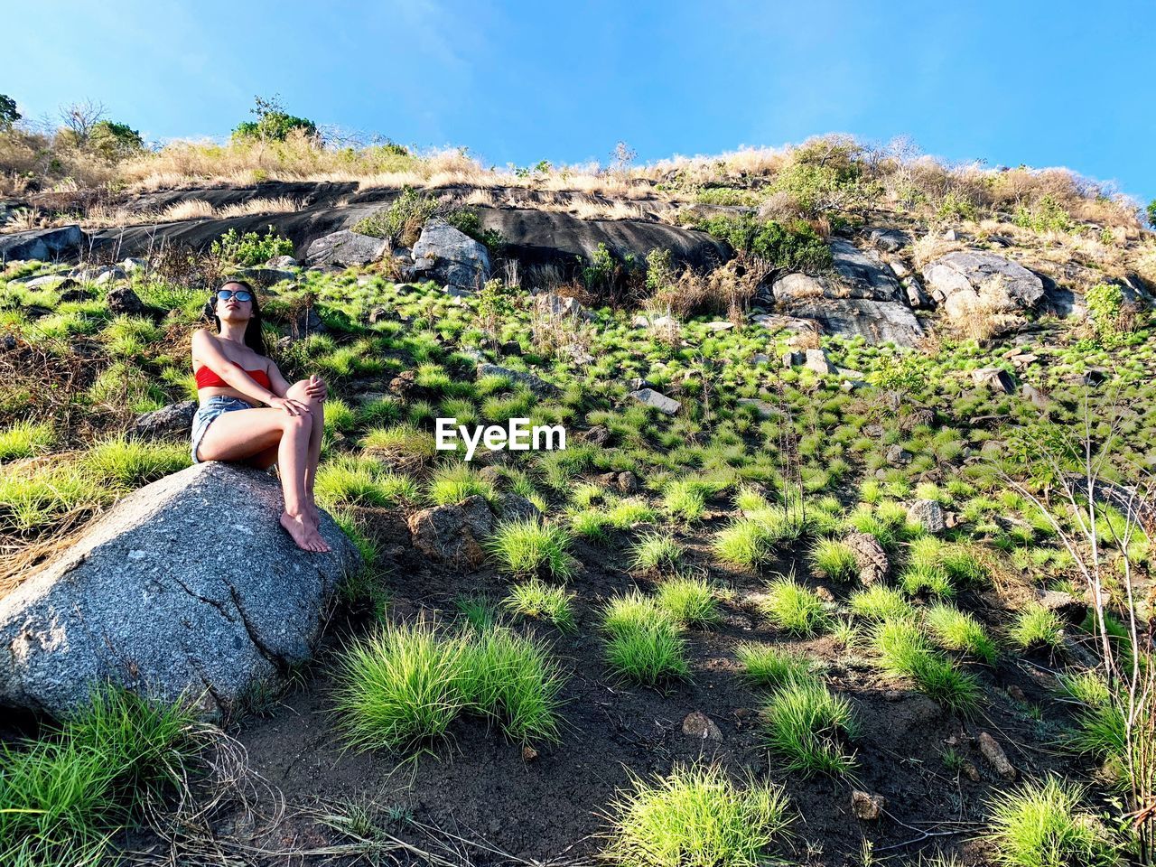 SIDE VIEW OF YOUNG WOMAN SITTING ON ROCK AGAINST PLANTS