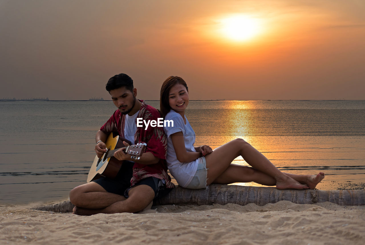 FRIENDS SITTING ON SHORE AT BEACH AGAINST SKY