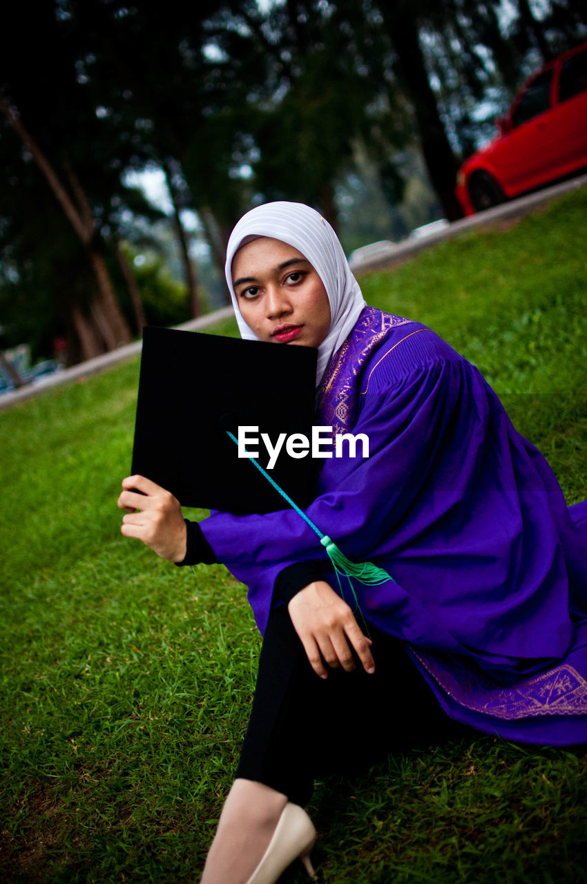 Portrait of young woman holding mortarboard while sitting on grassy land