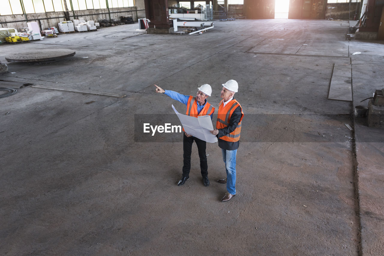 Two men with plan wearing safety vests talking in old industrial hall