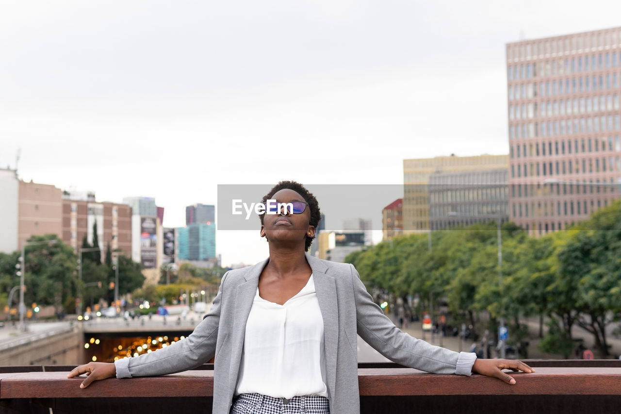 Young black female manager wearing trendy outfit and eyeglasses leaning on railing while resting with eyes closed in balcony of modern building