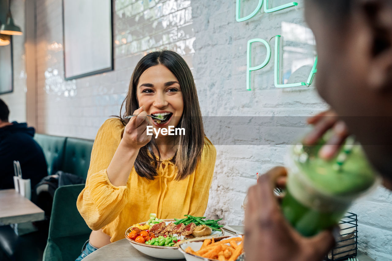 Content female enjoying tasty dish while interacting with crop unrecognizable ethnic partner drinking smoothie in restaurant