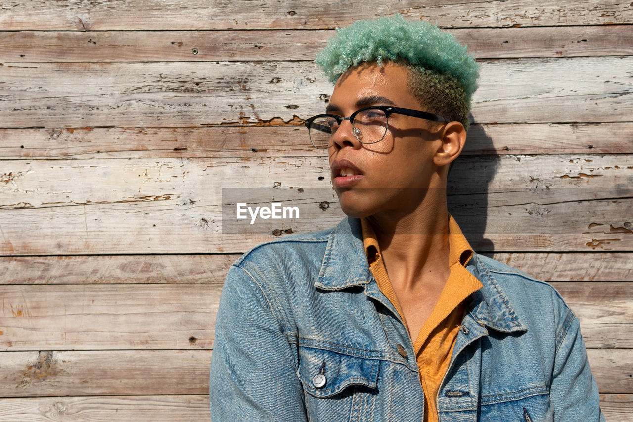 Optimistic black male hipster with blue curly hair standing near wooden building background on sunny day while looking away