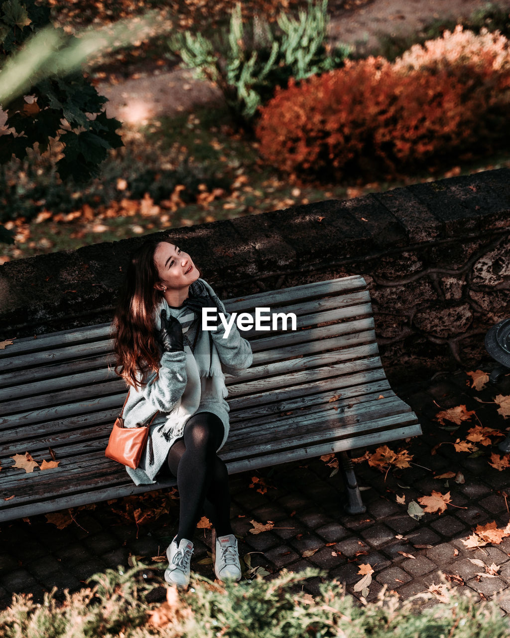 FULL LENGTH OF YOUNG WOMAN SITTING ON BENCH IN PARK