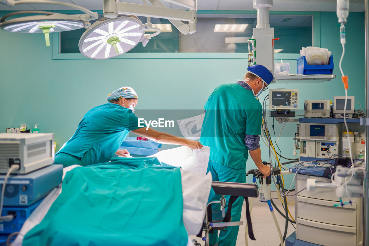 Back view of unrecognizable male and female medical assistants in uniforms preparing couch and devices in operating room for surgery in modern hospital