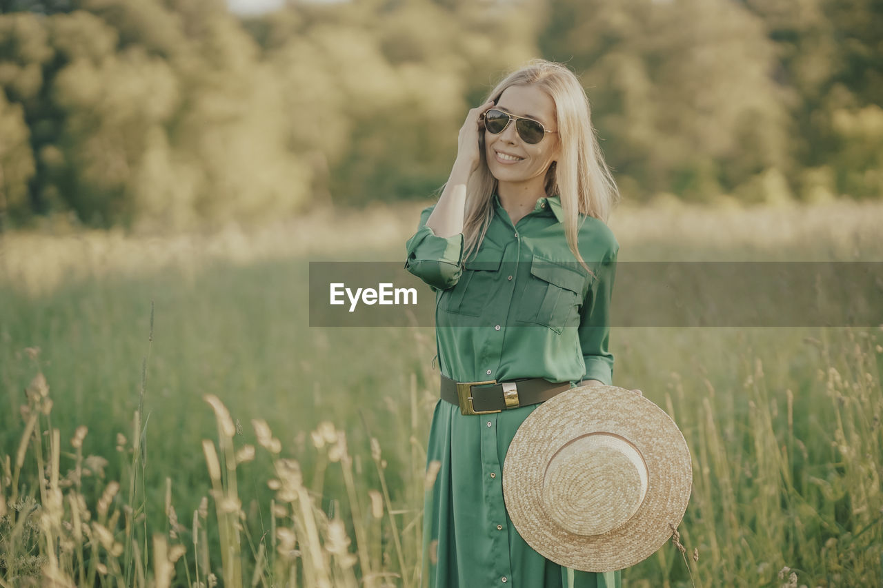 one person, women, plant, adult, grass, blond hair, field, nature, land, rural scene, landscape, clothing, long hair, agriculture, smiling, fashion, young adult, hat, green, portrait, hairstyle, happiness, emotion, person, summer, three quarter length, female, environment, portrait photography, outdoors, front view, lifestyles, day, leisure activity, beauty in nature, sky, standing, casual clothing, meadow, plain, dress, spring, glasses, crop, copy space, tranquility, sun hat, fashion accessory