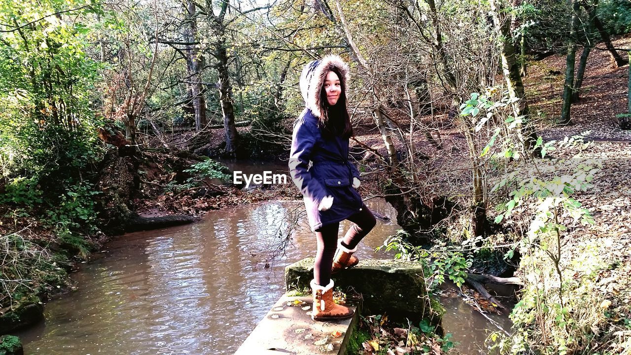 Full length portrait of smiling woman standing with hands in pockets by stream in forest
