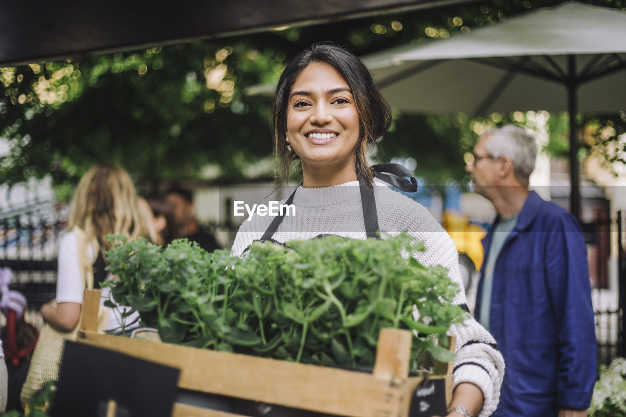 Young smiling female florist holding crate of plants at flea market