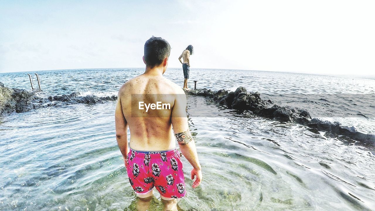 REAR VIEW OF SHIRTLESS MAN LOOKING AT SEA SHORE