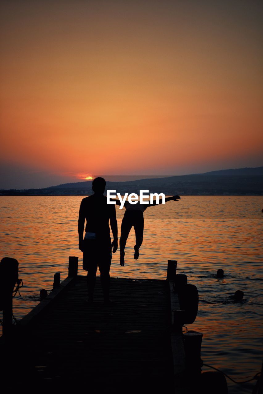 Rear view of man on pier looking at friend jumping into sea during sunset