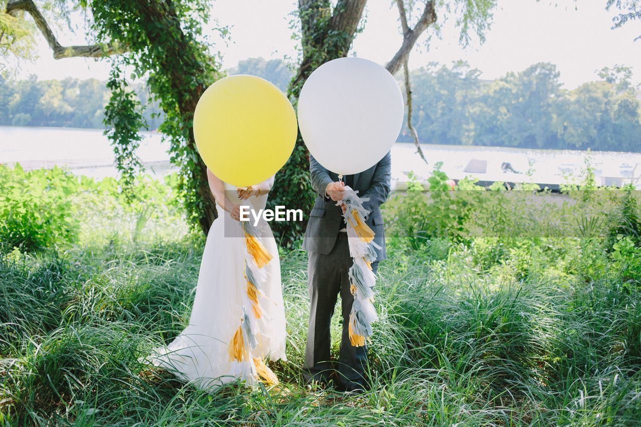 Bride and bridegroom standing with balloons in front of face on grass