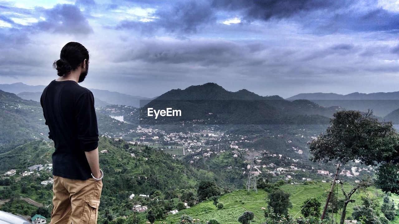 REAR VIEW OF MAN LOOKING AT MOUNTAIN RANGE AGAINST SKY