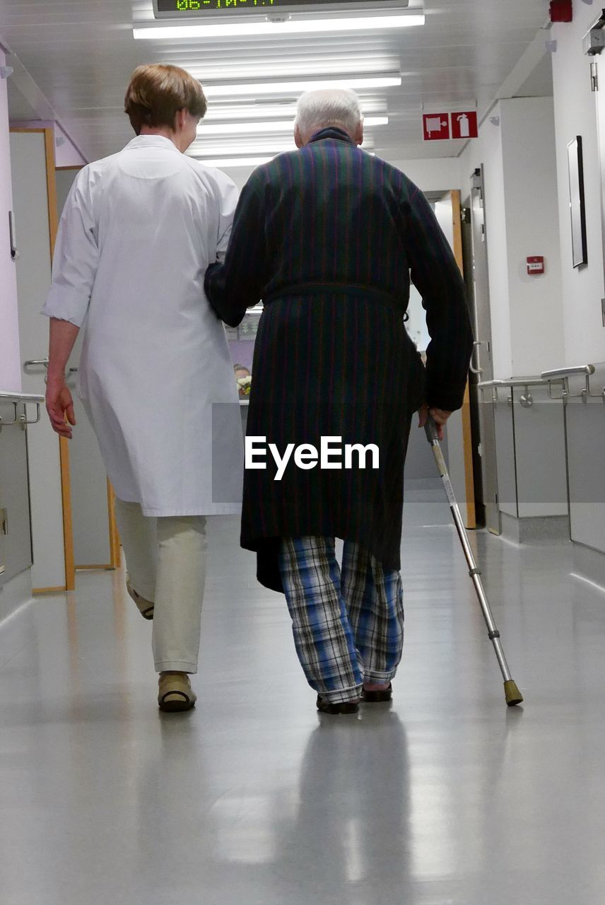 Rear view of senior man walking with nurse in hospital corridor