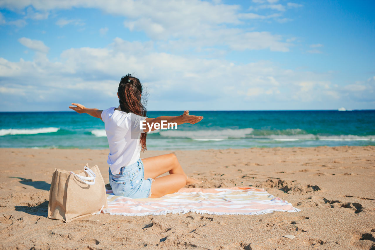 rear view of woman with arms outstretched standing at beach against sky