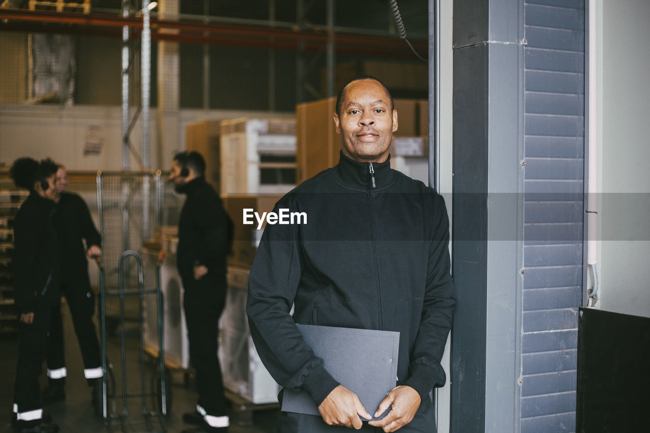 Confident male entrepreneur standing with clipboard at logistics warehouse