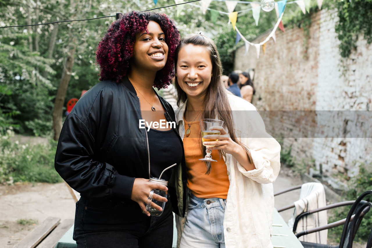 Portrait of happy multiracial female friends with drinks standing together during party in back yard