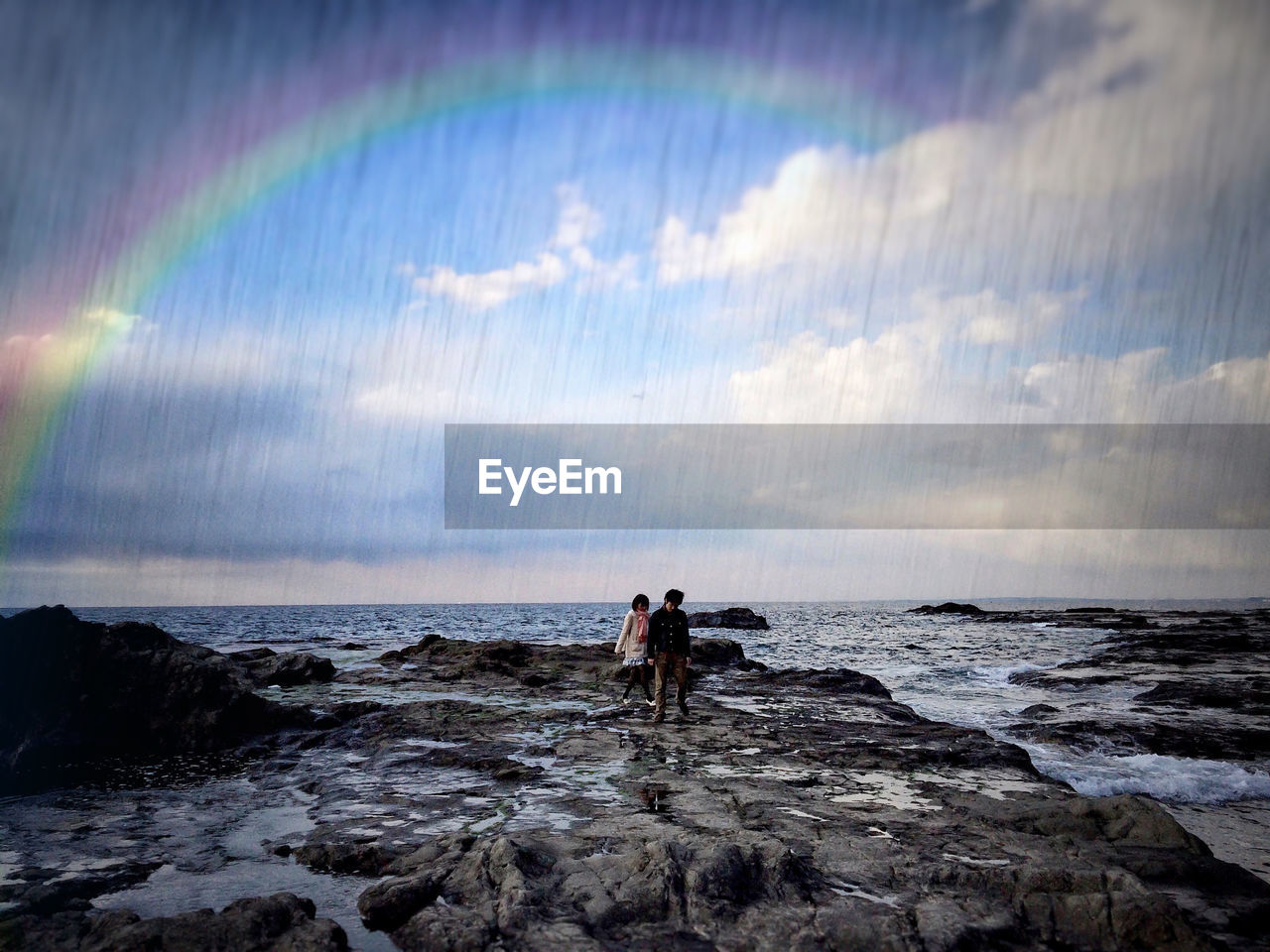People on rocky shore against rainbow in sky during monsoon