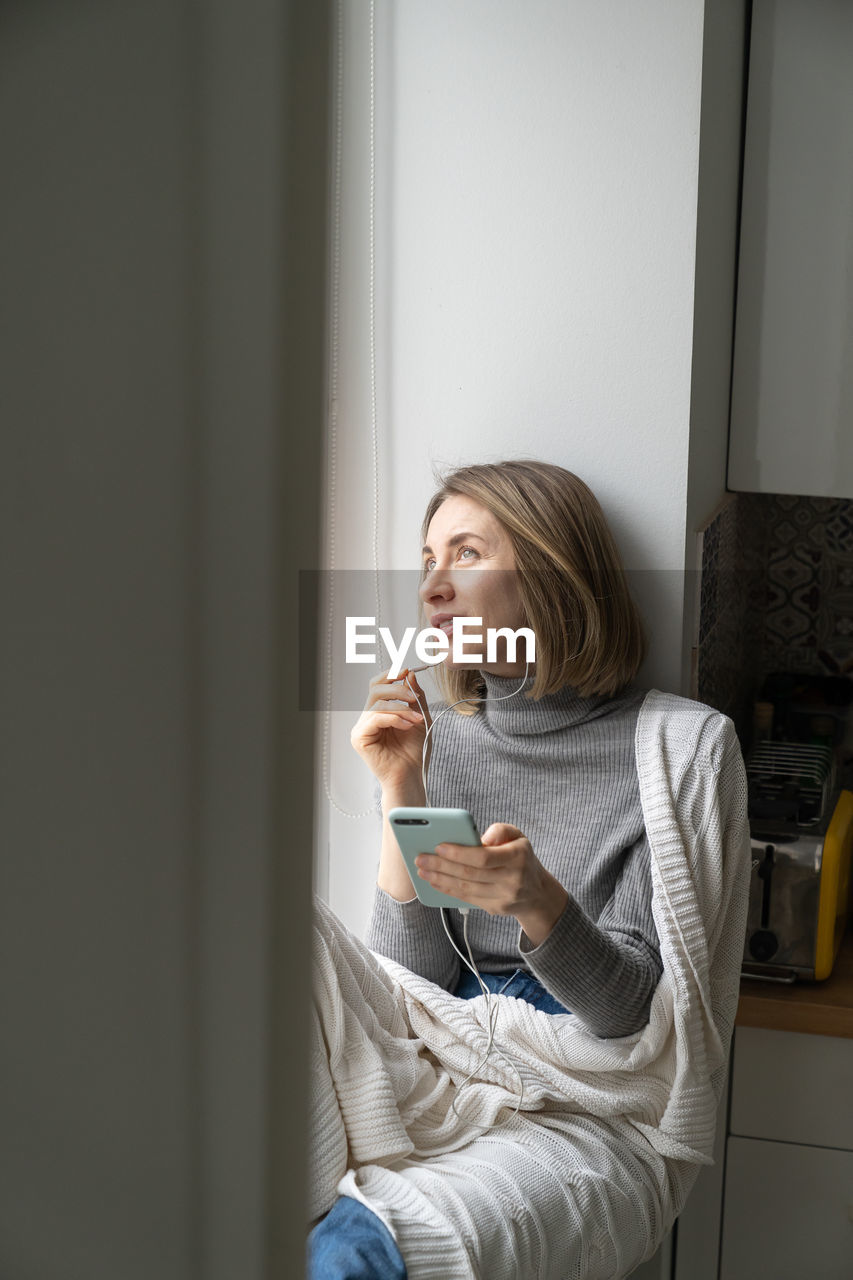 Smiling woman looking away while sitting by window with smart phone