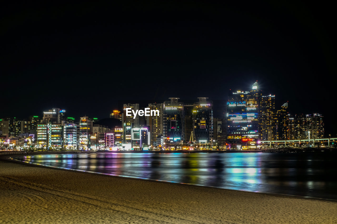 ILLUMINATED MODERN BUILDINGS BY RIVER AGAINST SKY AT NIGHT