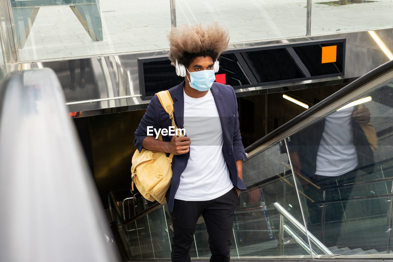 Young african man wearing a mask on an escalator with a bag hanging over his shoulder while listening to music with a wireless headset