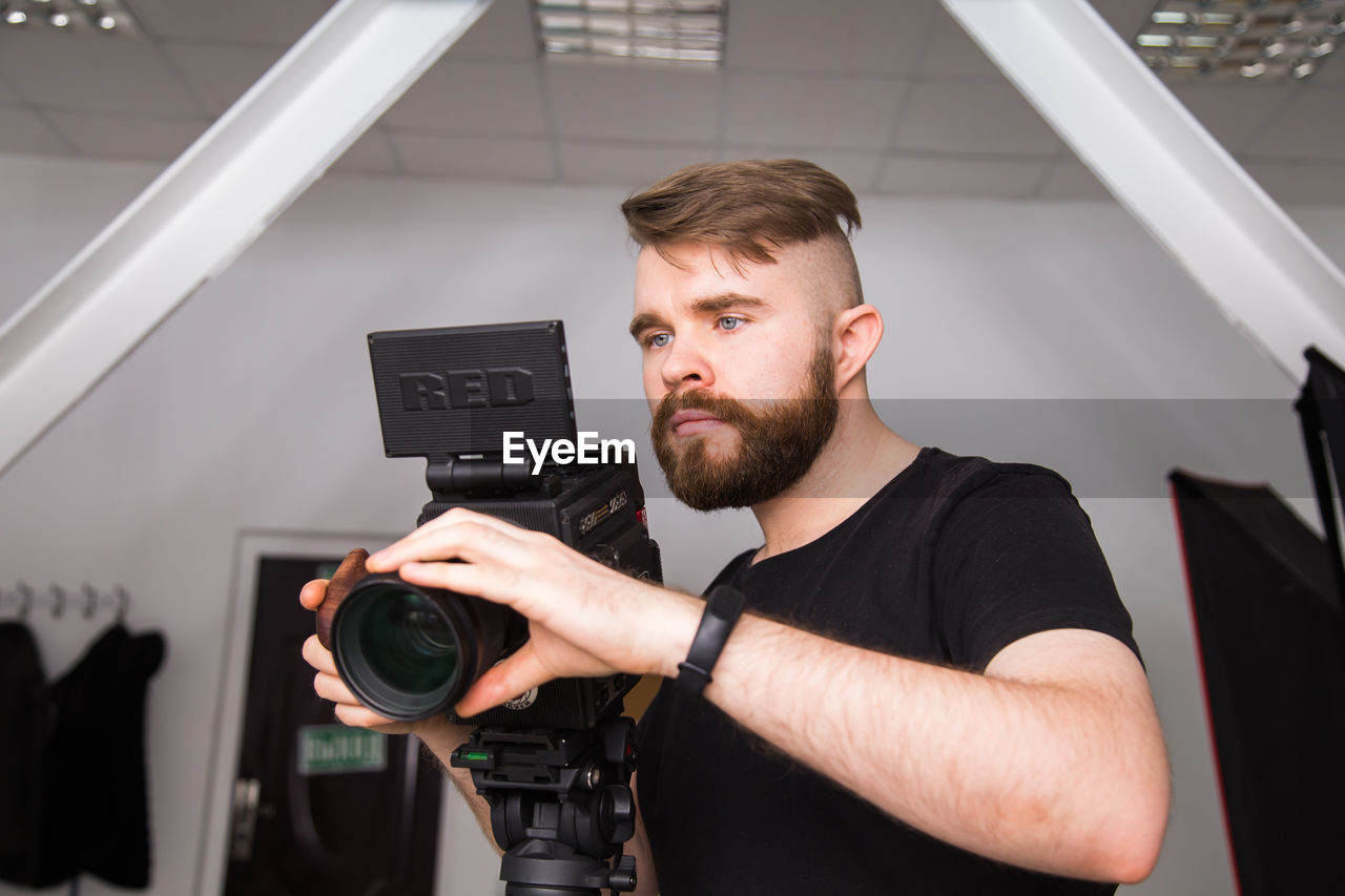 one person, adult, beard, young adult, occupation, indoors, facial hair, technology, men, arts culture and entertainment, camera, creativity, waist up, portrait, business, standing, holding, expertise, studio, looking, working, film industry, casual clothing, business finance and industry, front view