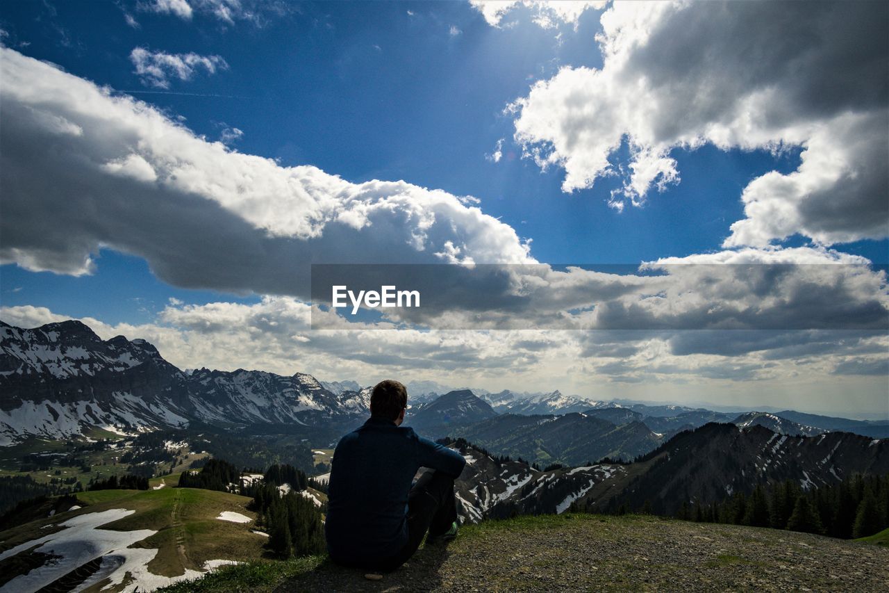 Rear view of man sitting on mountain against cloudy sky