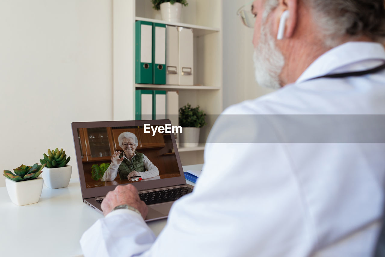 Back view of crop anonymous elderly male doctor in wireless earbud speaking with woman on video call on netbook in clinic