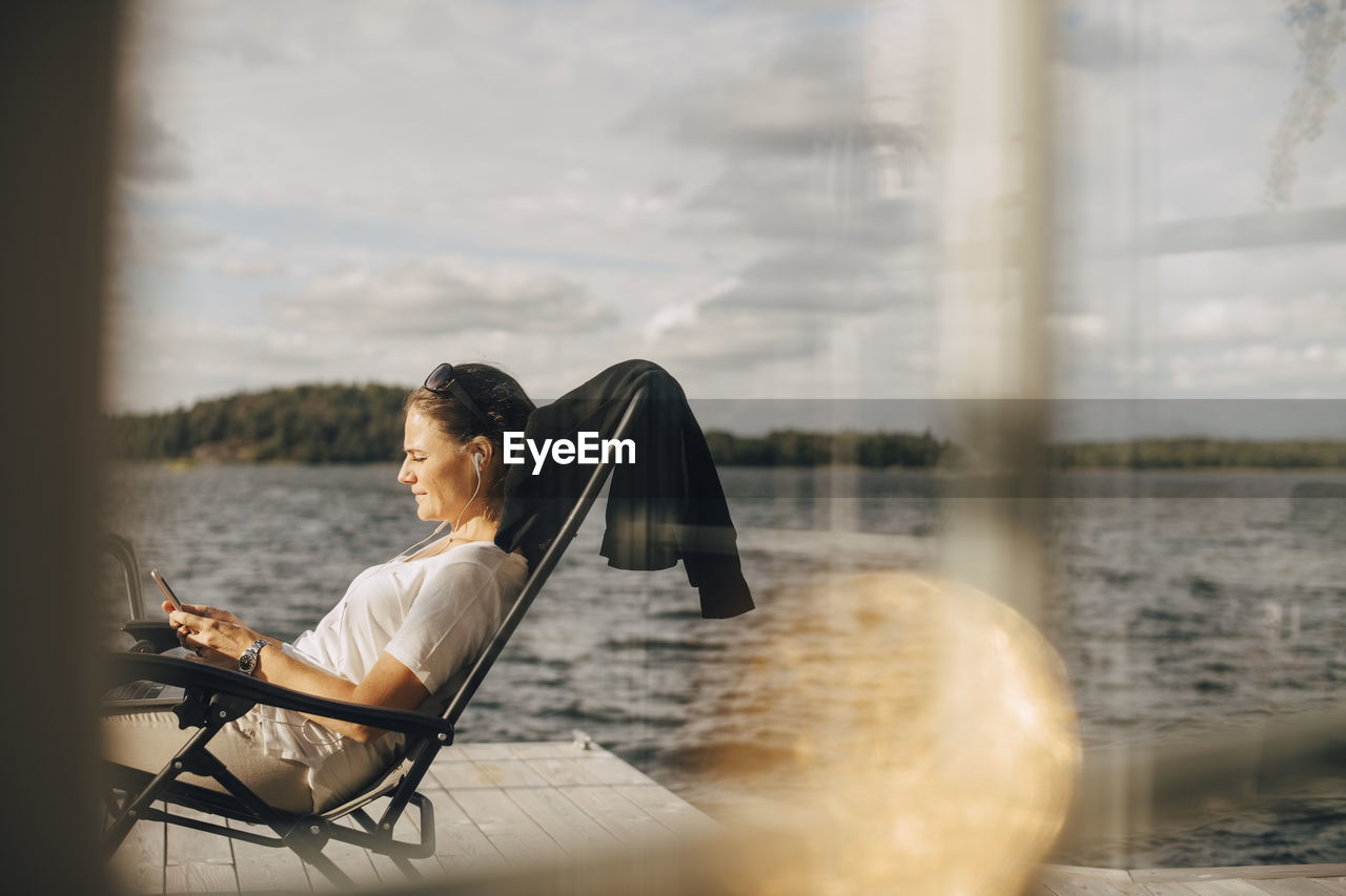 Side view of woman wearing headphones while using phone on patio seen through window