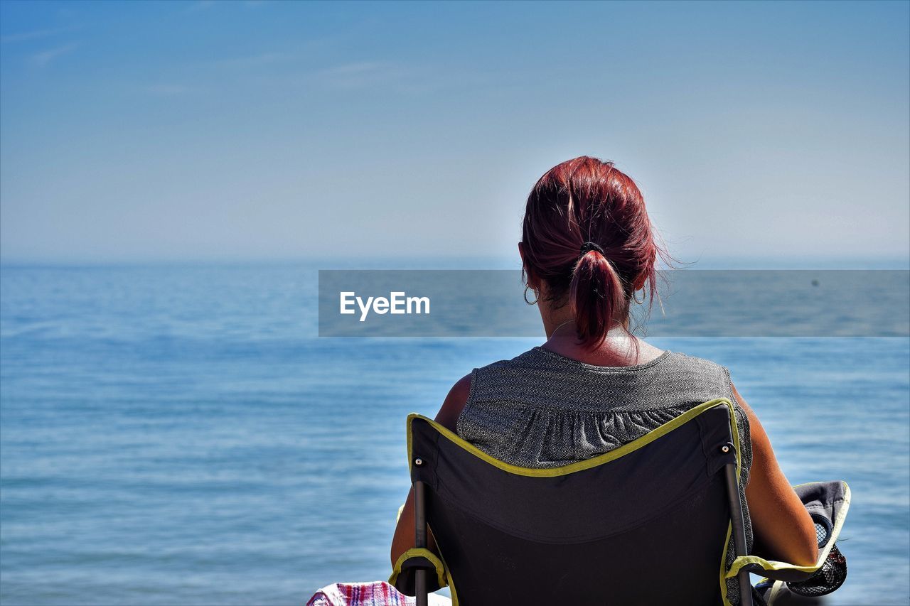 Rear view of mature woman relaxing on chair at beach against sky during sunny day